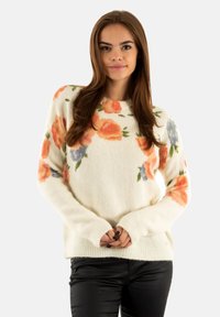 Joven con cabello largo y castaño, vistiendo un suéter blanco con patrones florales en naranja y azul, y pantalones negros, de pie con las manos entrelazadas.