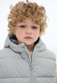 Jasnoszara kamizelka puchowa z kapturem, zamkiem błyskawicznym i quiltowanym wzorem, noszona na beżowym swetrze z dzianiny. Miękka tekstura, luźny krój.