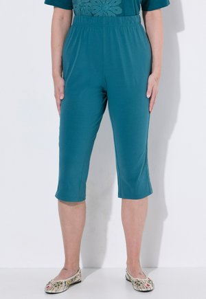 Kobieta w turkusowych krótkich spodniach z elastycznym pasem i balerinach w kwiatowy wzór, stojąca na białym tle.