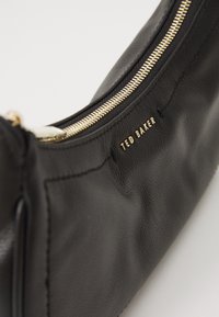Schwarze Lederhandtasche mit strukturierter Oberfläche und goldenem Reißverschluss. Verfügt über den eingeprägten Markennamen "TED BAKER" in der Nähe des Reißverschlusses. Gebogene Form.