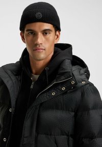 Giacca a vento nera con un motivo testurizzato, accessori in metallo argentato e cappuccio, indossata sopra un hoodie nero con zip e abbinata a un berretto nero.