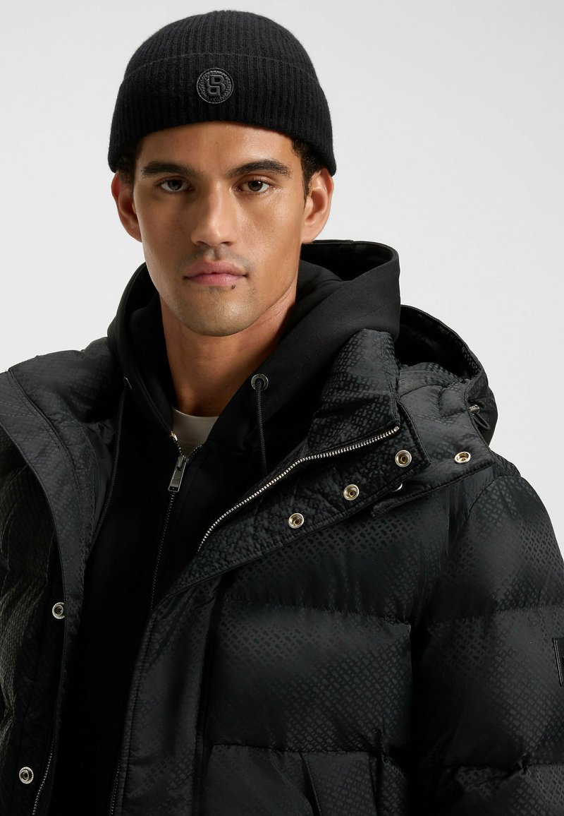 Giacca a vento nera con un motivo testurizzato, accessori in metallo argentato e cappuccio, indossata sopra un hoodie nero con zip e abbinata a un berretto nero.