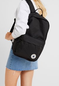 Sac à dos tote noir avec double bretelles, poche avant et écusson logo Converse. Fabriqué en tissu durable avec une texture lisse.