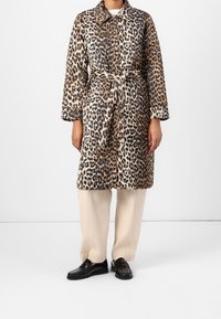 Personne portant un manteau ceinturé à imprimé léopard, un pantalon ample crème et des mocassins noirs, debout devant un fond blanc uni.