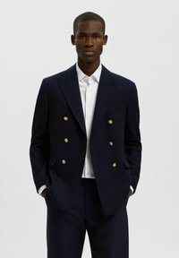 Granatowy dwurzędowy blezer z złotymi guzikami, dopasowany fason, klapy z wycięciem i boczne kieszenie, noszony na białej koszuli.