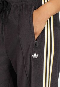 Black athletic joggers made of textured fabric with a zippered pocket on the left and three vertical stripes in yellow and white accents.