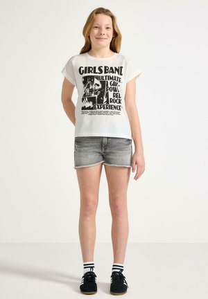 Ung pige stående iført en hvid t-shirt med "Girls Band" grafik, grå denimshorts, stribede sokker og sorte sneakers mod en hvid baggrund.