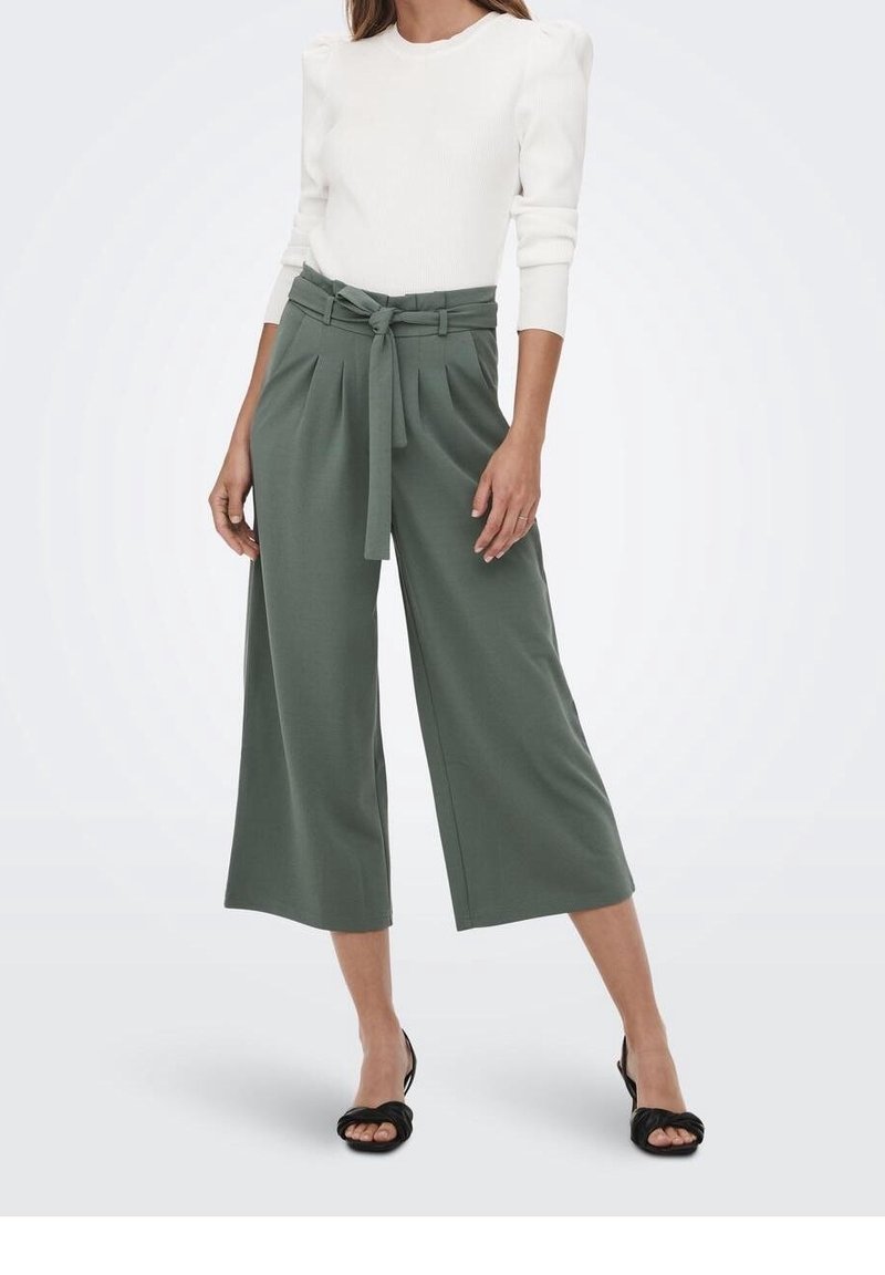 Vrouw in een witte lange mouw top en hoge taille wijde groene cropped broek met riem, gecombineerd met zwarte open teen sandalen.
