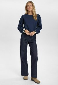 Felpa blu navy con polsini a righe, abbinata a jeans larghi scuri e stivaletti alla caviglia color sabbia. La felpa ha un collo rotondo e una vestibilità comoda.