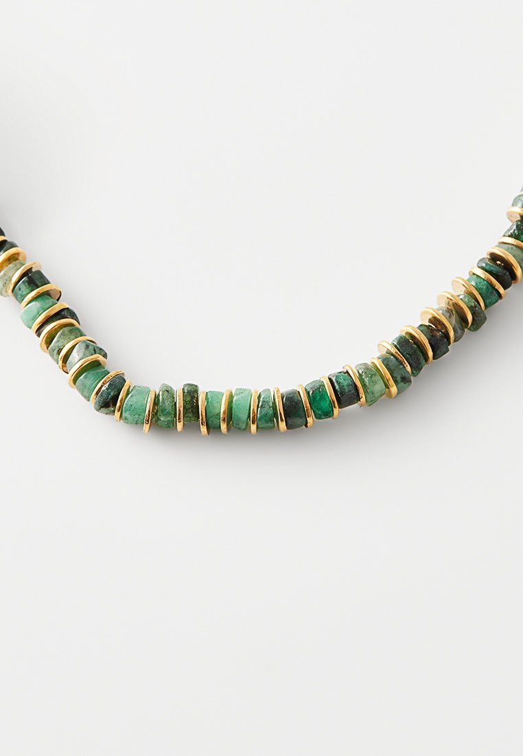 Collier avec des petites perles en pierre verte alternées et de fins disques en or espacés, disposés en ligne courbe sur un fond blanc.