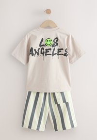Hellbeiges T-Shirt mit schwarzem "LOS ANGELES"-Aufdruck und grünem Smiley, kombiniert mit gestreiften Shorts in Weiß, Grau und neon-grünen Akzenten.