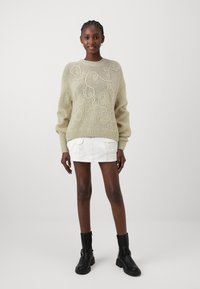 Maglione lavorato a maglia color beige chiaro con design astratto testurizzato, polsini e orlo a coste. Abbinato a una mini gonna bianca e stivaletti neri alla caviglia.