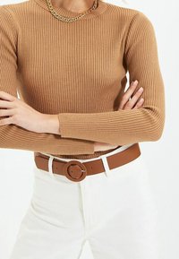 Haut marron à manches longues côtelé, associé à une ceinture en cuir marron avec une boucle ronde, porté par-dessus un pantalon blanc. Tissu texturé et coupe structurée.