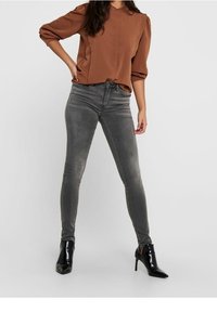 Braunfarbige Bluse mit Puffärmeln, kombiniert mit grauen Skinny-Jeans. Schwarze High-Heels. Minimalistisches Design mit glatten Texturen.