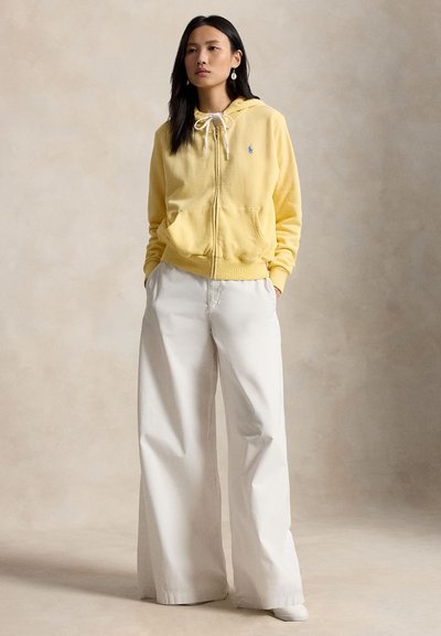 Femme debout, les mains dans les poches, portant un sweat à capuche jaune zippé et un pantalon blanc à jambes larges sur un fond neutre.