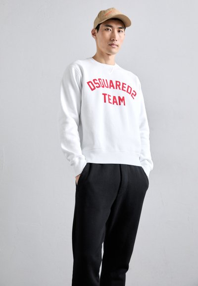Young man standing with hands in pockets wearing white sweatshirt with red "DSQUARED2 TEAM" text, black pants, and beige cap on neutral background.