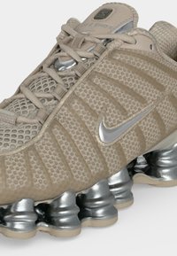 Pantof sport bej Nike Shox cu parte superioară din plasă, logo Nike argintiu, șireturi bej și unități metalice argintii pentru talpă cu pernă de aer vizibile.