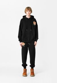 Sudadera negra con un parche de oso en el pecho, combinada con pantalones deportivos negros que tienen un parche de oso en el bolsillo. Botas marrones de cordones completan el conjunto.