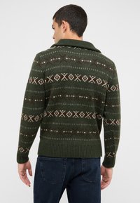 Zelený vzorovaný sveter s geometrickými motívmi v krémovej a hnedej farbe, s rebrovaným lemom a šálovým golierom, vyrobený z mäkkého pleteného materiálu.