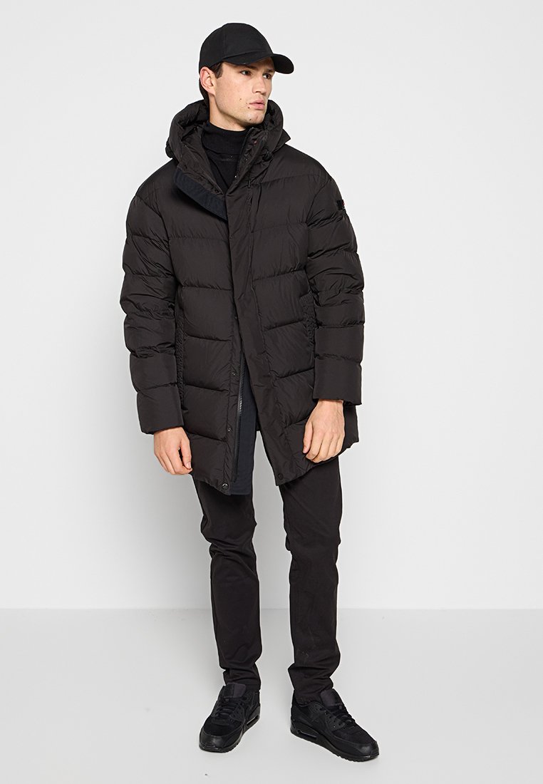 Zwarte pufferjas met een capuchon, met een gewatteerd ontwerp en een voorknoopsluiting. Draagt over een zwarte top en gecombineerd met zwarte broeken en schoenen.