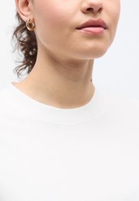 Χρυσό μεταλλικό σκουλαρίκι κρίκος με λεία, αντανακλαστική επιφάνεια· φορεμένο με απλό λευκό πουκάμισο. Εστίαση στην περιοχή του αυτιού και του λαιμού.