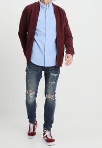 Muž kráčející v vínovém zipovaném kabátu, světle modré košili s knoflíky, potrhaných tmavomodrých džínách a červených teniskách s bílými tkaničkami.