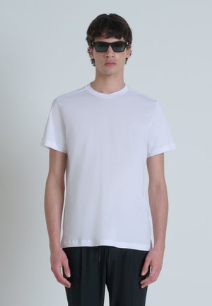 Camiseta blanca de manga corta en tejido suave, con cuello redondo y un logo sutil en el pecho. Combinada con pantalones negros.