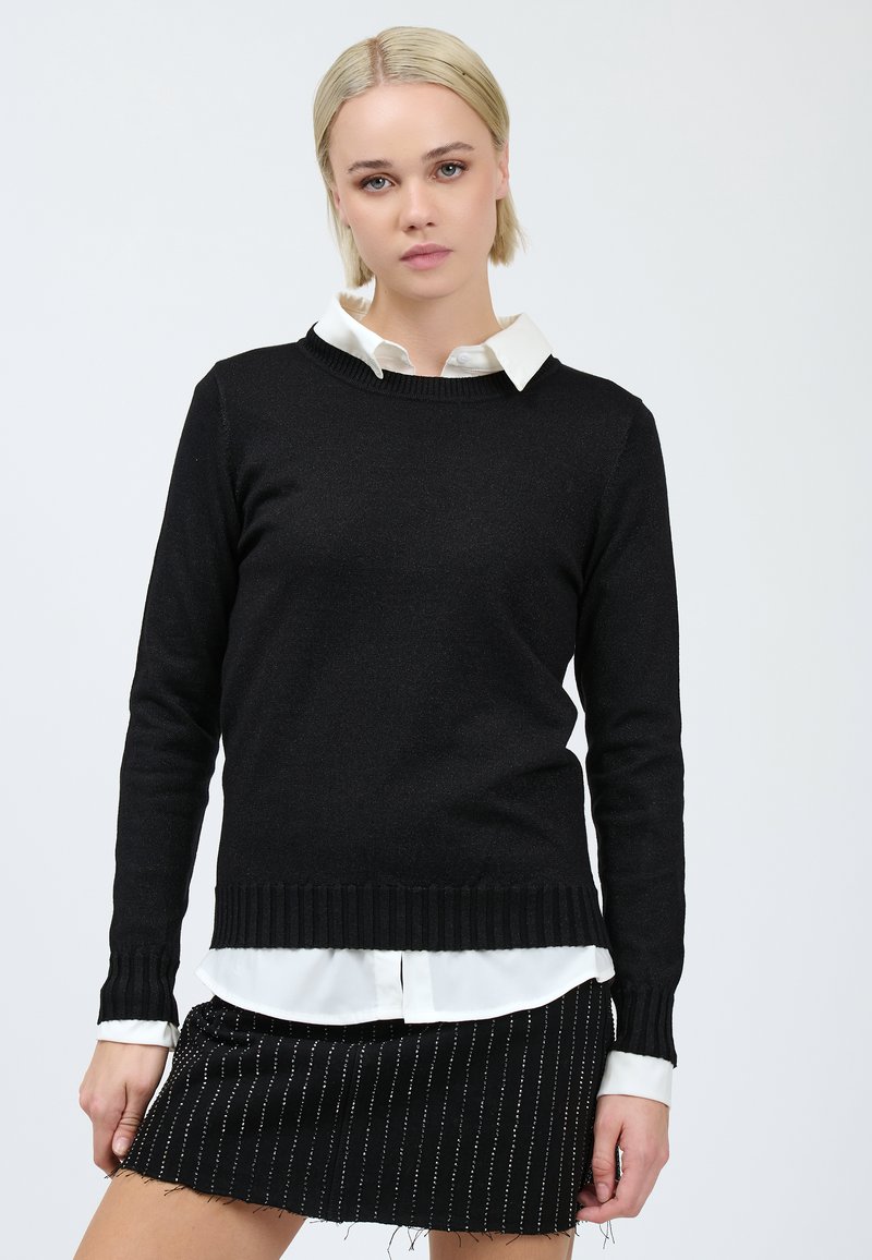 Una giovane donna con i capelli biondi corti indossa un maglione nero sopra una camicia bianca con colletto e una gonna nera a righe, in piedi davanti a uno sfondo neutro.