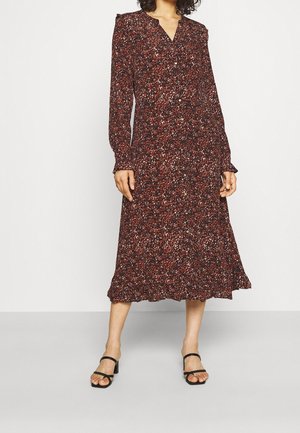 Femme portant une robe midi à manches longues boutonnée, fleurie rouge foncé, associée à des sandales à talons ouvertes noires sur un fond uni.