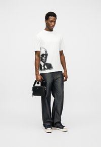 Άνδρας που φοράει λευκό μπλουζάκι με σχέδιο, σκούρο τζιν, μαύρα αθλητικά παπούτσια, κρατώντας μαύρη ορθογώνια τσάντα πάνω σε απλό φωτεινό φόντο.