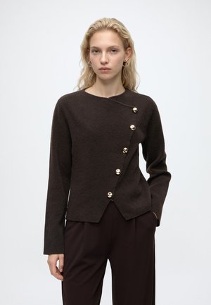 Jeune femme aux cheveux blonds portant un pull asymétrique boutonné marron foncé et un pantalon assorti sur un fond clair uni.