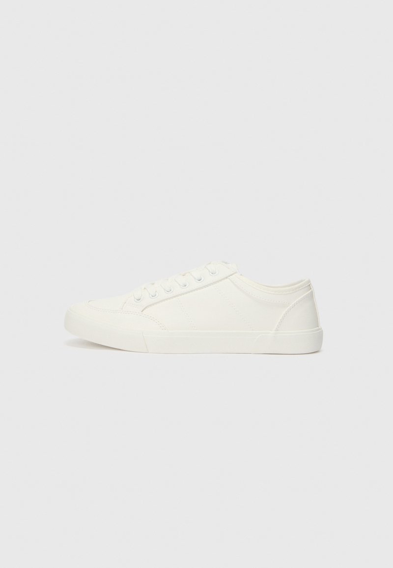 Baskets basses blanches en toile avec lacets blancs et semelle en caoutchouc, vues de profil sur un fond uni gris clair.