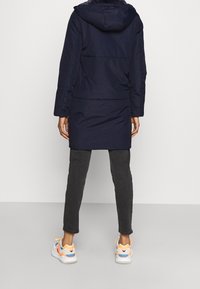 Mörkblå parka med huva, långa ärmar och en lätt A-linjeform. Bärs med mörkgrå byxor och färgglada sneakers. Slät tygerstruktur.