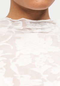 Κοντινό πλάνο ενός ατόμου που φορά λευκό διαφανές κορυφαίο με λουλουδάτη δαντέλα και ψηλό λαιμό, με φόντο ένα απλό ανοιχτόχρωμο τοπίο.