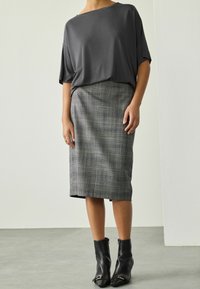 Eine Frau trägt ein lockeres, dunkelgraues Oberteil, einen knielangen grauen karierten Rock und schwarze Stiefeletten mit spitzen Zehen und Schnallen.