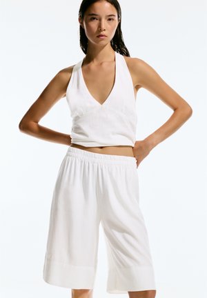 Hvid halter top med dyb V-udskæring og tætsiddende skød, parret med løse, knælange hvide shorts med elastisk talje.