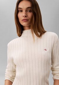 Jeune femme aux cheveux bruns et raides portant un pull côtelé crème avec un petit logo Calvin Klein rouge et blanc sur la poitrine.