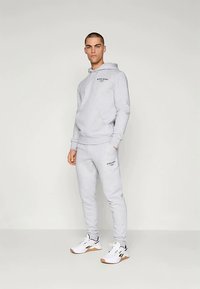 Γκρι φούτερ με κουκούλα και λογότυπο, σε συνδυασμό με αντίστοιχες φόρμες, και οι δύο με ελαστική ζώνη στη μέση. Λευκά αθλητικά παπούτσια με μαύρες λεπτομέρειες.