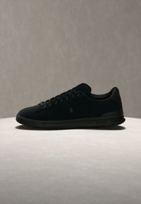 Μαύρο sneaker χαμηλής κοπής με κορδόνια, ένα διακριτικό λογότυπο στο πλάι και υφασμένο πέλμα, παρουσιασμένο σε ανοιχτή επιφάνεια με απλό φόντο.