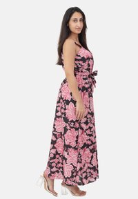 Donna in un maxi abito senza maniche nero con stampa floreale rosa e cintura in vita, che indossa sandali con tacco a blocco trasparenti, in posa di profilo.