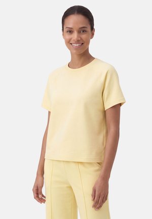 Kvinde smilende iført en bleg gul kortærmet top og matchende blege gule bukser mod en ensfarvet lys baggrund.