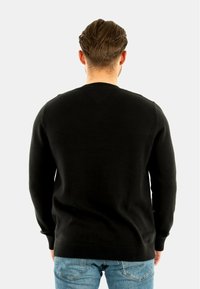 Svart stickad tröja med rundad halsringning, ribbade ärmslut och avslut, visad bakifrån. Combinerad med ljusblå denimjeans.