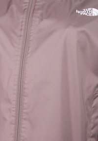 Lichtroze jas van gladde stof met een rits aan de voorkant. Voorzien van een wit logo aan de rechterbovenkant en een strak, minimalistisch ontwerp.