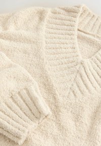 Krémový svetr s měkkou, texturovanou tkaninou. Má pružný límec a manžety, s designem do V a plyšovým povrchem.