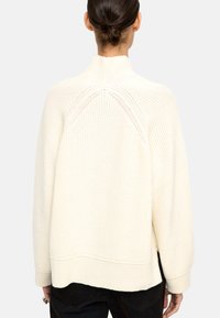 Kremowy sweter z dzianiny o wysokim kołnierzu, prążkowanej teksturze, luźnym kroju i opadających ramionach. Posiada rozcięcia po bokach u dołu.