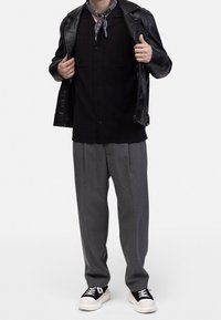 Zwarte leren jas over een zwart overhemd met knopen, grijze geplooide broek en zwarte sneakers met witte zolen. Bandana om de hals.