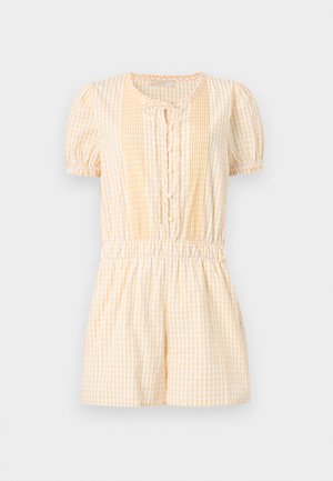 Naine, kes kannab kollase ja valge triibulise lühikeste varrukatega kombinesooni, millel on nööbid ja elastne vöökoht, seisab käed taskutes ja mustade sandaalidega.