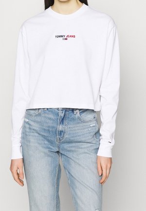 Kobieta ubrana w białą, długą, skróconą bluzkę z logo „TOMMY JEANS” oraz jasnoniebieskie, wysokie jeansy, stojąca na jednolitym tle.