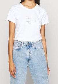 Bílý bavlněný tričko se malým zlatým logem "Tommy Jeans" na přední straně, spárované se světle modrými džíny s vysokým pasem.