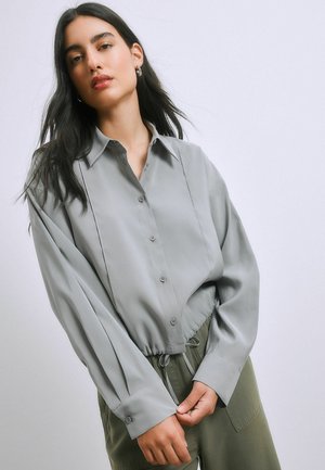 Frau mit langen dunklen Haaren, die ein locker sitzendes graues Hemd mit Knöpfen und Kordelzug in der Taille sowie olivgrüne Hosen trägt, vor einem einfachen Hintergrund.